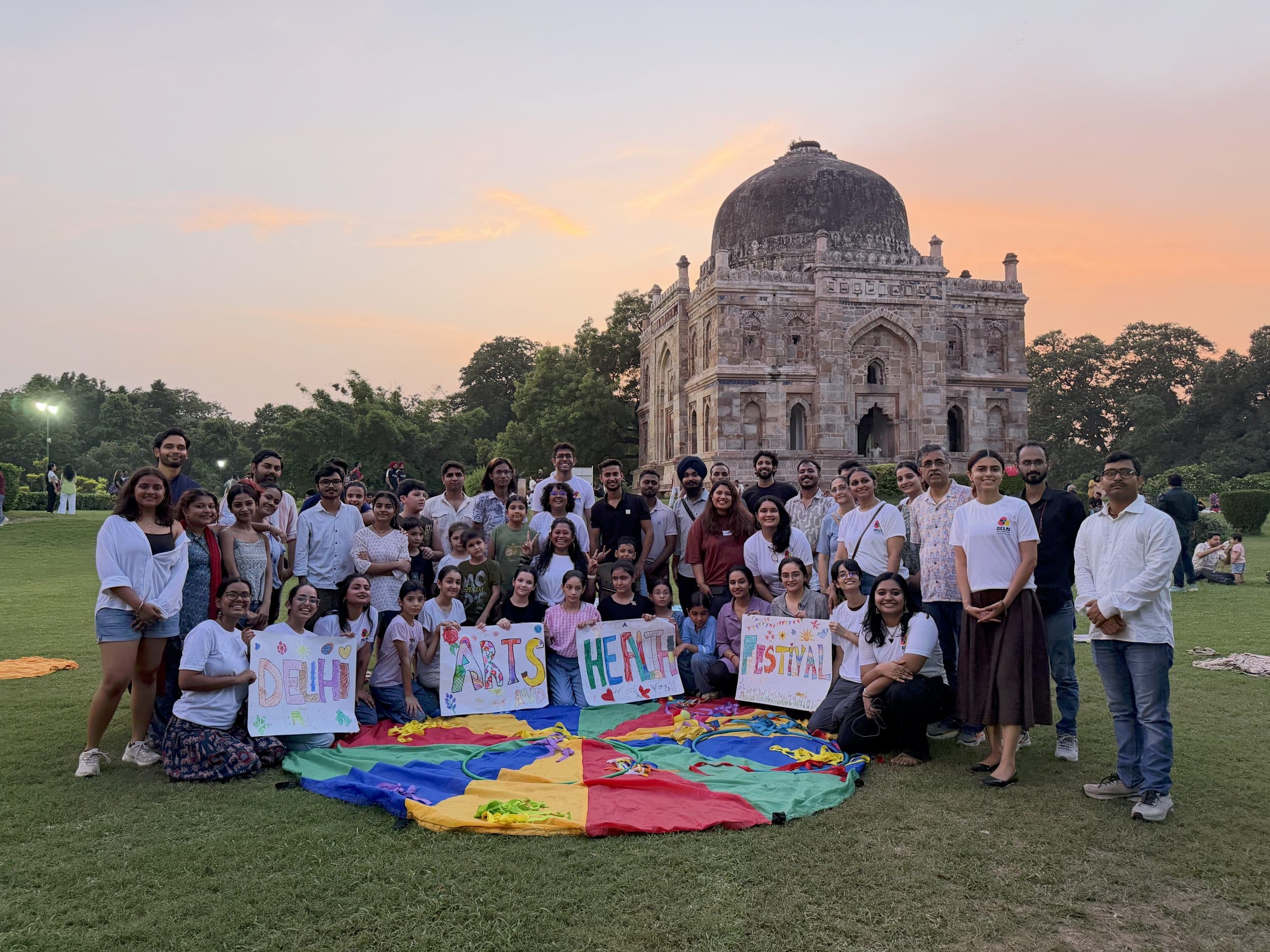 A large group holding DELHI, ARTS, HEALTH and FESTIVAL signs together on the grass at sunset in front of a domed tomb